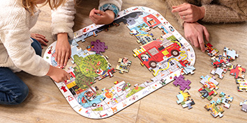 Zwei Personen setzen ein buntes Puzzle von My Mandarine auf einem Holzboden zusammen. Das Puzzle zeigt ein Feuerwehrauto und verschiedene Comic-Elemente. Um den teilweise fertigen Puzzlerahmen herum liegen verstreute lose Teile.