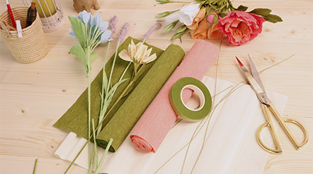 Grüne und rosafarbene Rolle Krepppapier von Clairefontaine auf hellbraunem Holztisch liegend. Zusammen mit einer Schere, Kreppband, Kunstblumen und Stiften.