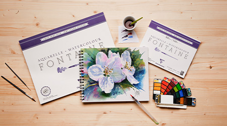 Ein Aquarellset auf einem Holztisch mit offener Farbpalette, Pinseln und Papierblöcken mit der Aufschrift „Aquarelle Fontaine“, ein Aquarellpapier von Clairefontaine. In der Mitte befindet sich ein fertiges Gemälde weißer Blumen auf einem Spiralskizzenbuch.