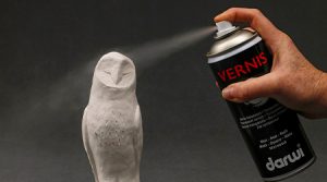 Eine Hand sprüht Klarlack von Darwi aus einer Dose auf eine weiße Eulenskulptur vor dunklem Hintergrund. Der Sprühnebel erzeugt einen Nebel über der Skulptur.