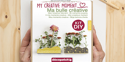Nahaufnahme von Händen, die eine DIY-Kit-Box mit der Aufschrift „Mein kreativer Moment“ halten. Die Box von Décopatch zeigt Bilder von Pflanzen, die in zwei dekorativen Haltern wachsen. Der Hintergrund ist eine Holzoberfläche. Auf der Verpackung sind verschiedene Sprachen zu finden.