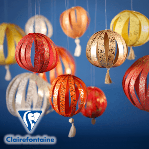 Bunte Papierlaternen mit Quasten hängen vor einem blauen Hintergrund in leuchtenden Rot-, Orange-, Gelb- und Beigetönen. Das Clairefontaine-Logo erscheint in der unteren linken Ecke und unterstreicht das Angebot an kreativem Kunstbedarf und Büroaccessoires von Exaclair.