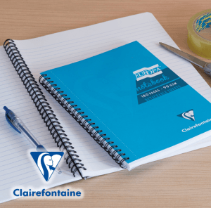 Ein blaues Clairefontaine-Spiralheft und ein Stift liegen auf einem aufgeschlagenen linierten Notizbuch, daneben liegen Schere und Klebeband. Büroaccessoires von Exacompta vervollständigen diesen stilvollen Arbeitsplatz auf einem Holzschreibtisch.