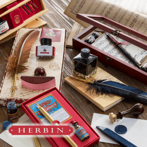 Eine Sammlung von Herbin-Schreibsets auf einem Holztisch mit Exacompta Clairefontaine-Tintenflaschen, Federkielen, Siegellack, Stempeln und Umschlägen. Im Hintergrund sind Notenblätter und elegante Verpackungen zu sehen.