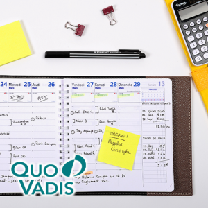 Ein offener Planer mit handschriftlichen Notizen, ein Stift, gelbe Haftnotizen, Heftklammern und ein Taschenrechner sind auf einer weißen Fläche angeordnet. Das Quo Vadis-Logo erscheint in der unteren linken Ecke und unterstreicht die Qualität von Exacompta Clairefontaine.
