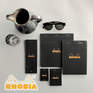 Ein flaches Foto mit verschiedenen schwarzen Rhodia-Notizblöcken, einer Sonnenbrille, einem Keramikkrug, einem Exacompta Clairefontaine-Büroklammerhalter aus Metall und einem großen Rhodia-Logo in der unteren linken Ecke auf hellgrauem Hintergrund.