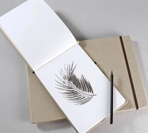 Ein aufgeschlagenes Skizzenbuch mit einer Schwarz-Weiß-Zeichnung eines Farnblattes liegt auf einem geschlossenen beigen Notizbuch, daneben ein schwarzer Bleistift auf einer hellgrauen Fläche.