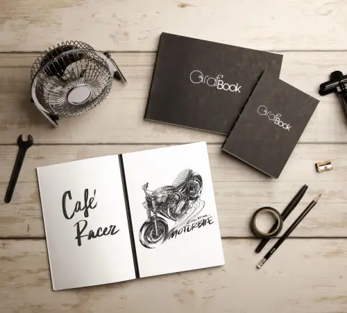 Ein aufgeschlagenes Skizzenbuch mit einer Café-Racer-Motorradzeichnung und handgeschriebenem Text, umgeben von einem weiteren geschlossenen Skizzenbuch, einem Fächer, einer Kamera, einem Schraubenschlüssel, Bleistiften, Klebeband und einem Spitzer auf einem Holztisch.