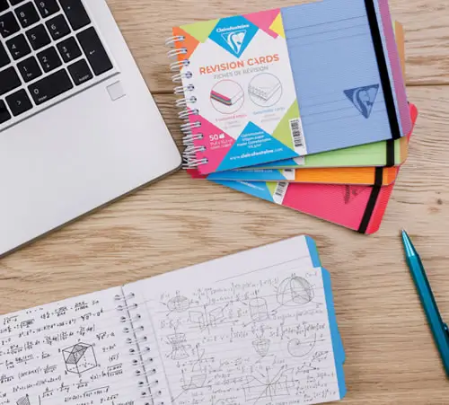 Auf einem hölzernen Schreibtisch liegen ein Laptop, bunte Lernkarten und ein aufgeschlagenes Notizbuch mit komplexen mathematischen Diagrammen und Gleichungen, daneben ein blauer Stift.