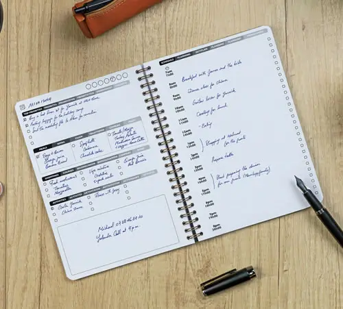 Ein offener, spiralgebundener Planer mit handschriftlichen Notizen und einem Zeitplan auf einem Holztisch, daneben ein schwarzer Stift und ein braunes Federmäppchen.