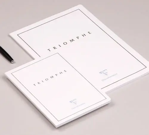 Zwei weiße Schreibblöcke mit der Aufschrift Triomphe und minimalistischen schwarzen Rändern, ein größerer und ein kleinerer, liegen nebeneinander auf einer hellen Fläche. Ein schwarzer Stift ist teilweise daneben sichtbar.