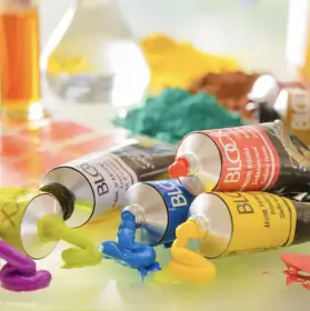 Drei Tuben mit leuchtender Acrylfarbe - grün, blau und gelb - spritzen bunte Acrylfarben auf eine Fläche, mit unscharfem Kunstzubehör und mehr Farbe im unscharfen Hintergrund.
