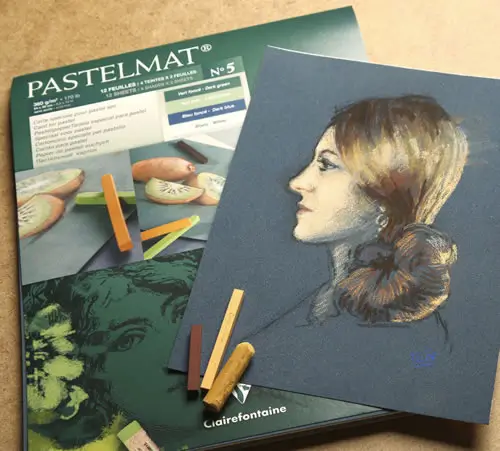 Eine Pastellzeichnung eines Frauenprofils liegt auf einem Pastellmatte-Papierblock, in dessen Nähe mehrere Pastellstifte verstreut sind. Der Block zeigt Abbildungen von Früchten und Produktangaben.